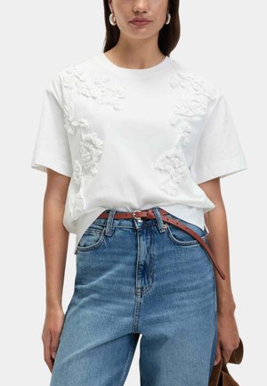 Weißes Baumwoll-T-Shirt mit strukturierten floralen Applikationsdesigns, kombiniert mit hochgeschnittenen blauen Jeans und einem braunen Gürtel.