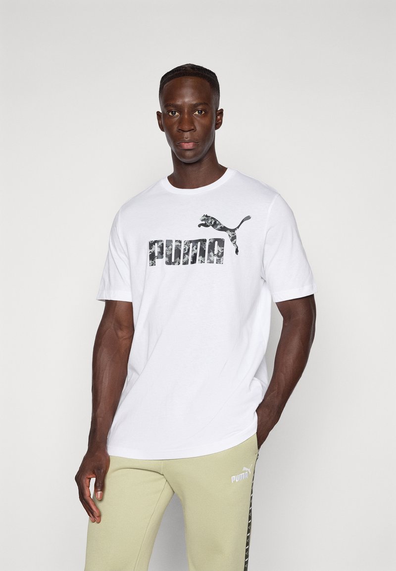 Vyras, vilkintis baltą Puma marškinėlį su kamufliažo logotipu ir šviesiai žaliais Puma sportiniais kelniais, prieš paprastą fono, pozuoja su viena ranka kišenėje.
