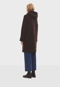Manteau à capuche marron en tissu texturé, à manches longues et à coupe décontractée, associé à un pantalon large bleu et des bottines cheville marron foncé.