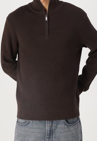 Hnědý zipovací svetr s texturovaným, vaflovým pletením. Má vysoký límec a dlouhé rukávy, kombinovaný se světle modrými džíny.