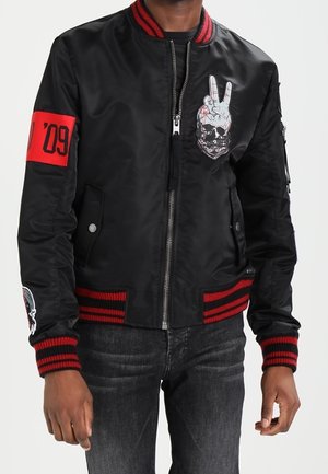 Personne portant une veste bomber noire avec des rayures rouges, un patch tête de mort et signe de paix sur la poitrine, et un patch rouge "09" sur la manche.