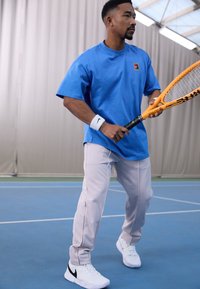 Blå bomull t-skjorte med en liten logo, lysegrå treningbukser med sidelister, og hvite joggesko med svart swoosh, holder en tennisracket.