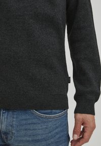 Pulover tricotat de culoare gri închis, cu manșete și tiv ribate, realizat dintr-un material moale, asortat cu blugi din denim albastru. Etichetă mică vizibilă pe lateral.