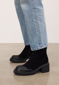 Mustad mustad ankle-bootid, paksude kontside ja tekstuursete kummist varvakaantega, kantud helesiniste denim teksadega betoonpinnal.