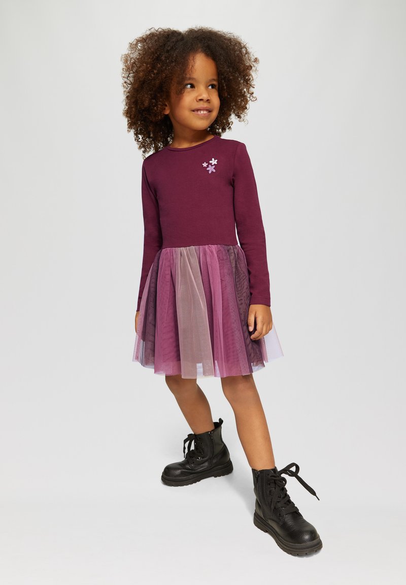 Robe violette à manches longues avec une jupe en tulle dans des nuances de rose et de lavande, ornée d'appliqués floraux. Associée à des bottes noires à lacets.