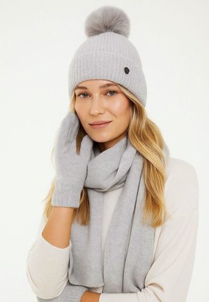 Bonnet en tricot côtelé gris avec un pompon, associé à une écharpe et des gants sans doigts assortis ; texture douce, design simple et détail de logo discret.