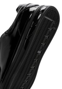 Zapatilla negra de cuero con acabado brillante, que presenta una suela de goma texturizada y un patrón en zigzag para mayor tracción. Diseño elegante y aerodinámico.