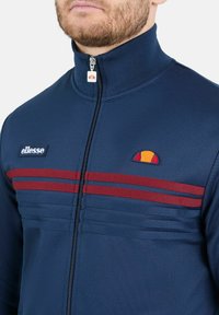 Veste zippée marine avec col haut, dotée de deux bandes horizontales rouges et d'un logo brodé sur la poitrine. Texture lisse.