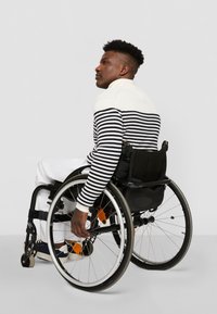 Gestreepte zwart-witte trui met hoge kraag, gedragen door een persoon in een rolstoel. Lichte broek en zwarte rolstoel met grijze wielen.