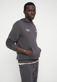 Hoodie cinza com um bolso frontal, tecido texturizado e detalhes bordados. Camada sobre uma camisa de cor clara e usada com calças de fato combinando.