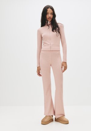 Ensemble en tricot rose clair composé de deux pièces avec un pull à col roulé zippé et un pantalon évasé, présentant un ourlet côtelé et une texture lisse. Des chaussures à plateforme viennent parfaire le look.