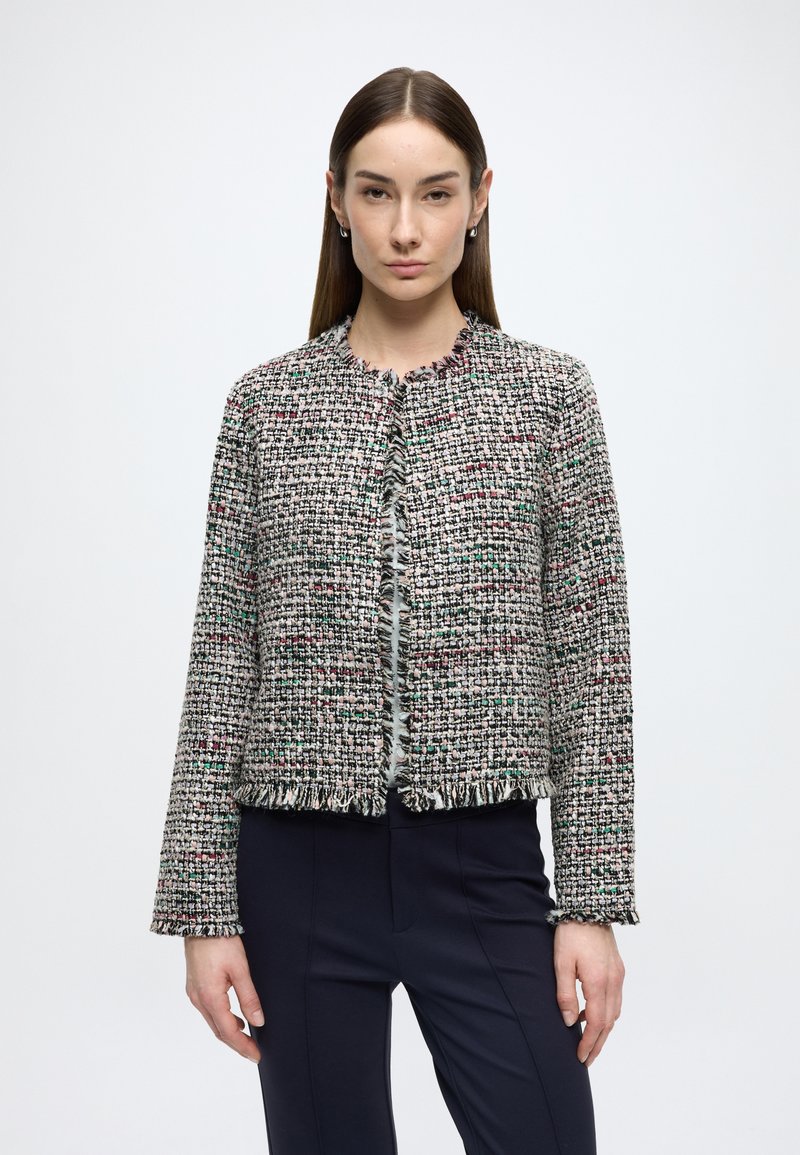 Jachetă țesută, texturată, în fire multicolore, cu decolteu rotund, tiv cu franjuri și mâneci lungi, purtată peste pantaloni închiși la culoare.
