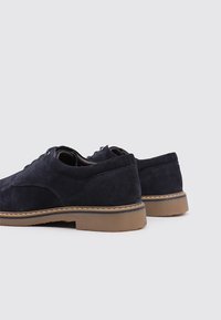 Zapatos de ante azul marino con un diseño de cordones, que cuentan con una suela de goma marrón clara y costuras en contraste a lo largo de los bordes.