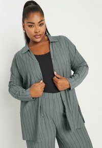 Cardigan a coste grigio chiaro con frontale a bottoni, maniche lunghe e vestibilità comoda, indossato sopra una canotta nera. La texture è morbida e lavorata.
