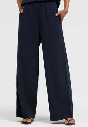 Person trägt locker sitzende marineblaue Weite-Hose mit elastischem Bund und Seitenschlitzen, Hände in den Taschen und schwarze Sandalen.