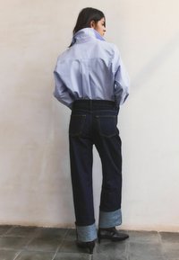 Mujer de espaldas con camisa azul claro con cuello levantado, jeans oscuros con puños y botines oscuros de tacón, sobre un suelo de baldosas.