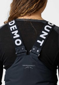 Spalline con testo bianco "DEMO" e "OUNT" indossate sopra una maglietta nera, attaccate a un capo nero con il logo "SWEDEMOUNT" e il profilo di una montagna.