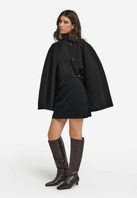 Schwarzer Umhang mit runden Knöpfen, getragen über einem schwarzen Rollkragenpullover und Rock. Braune kniehohe Lederstiefel mit spitzer Spitze und niedrigem Absatz.