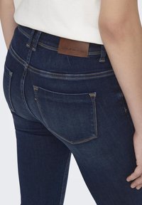 Jeans de mezclilla azul oscuro con un diseño ajustado. Presentan un parche de marca de cuero marrón en la parte trasera y dos bolsillos traseros con costura contrastante.