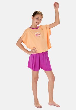 Jeune fille debout pieds nus, une main sur la hanche et l'autre levée, portant un t-shirt pêche et un short violet sur fond blanc.