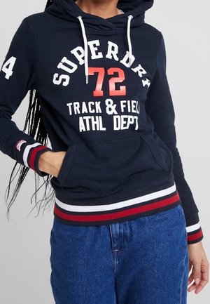 Osoba nosząca granatową bluzę z kapturem Superdry z czerwonym numerem „72” i białym napisem „TRACK & FIELD ATHL DEPT”, zestawioną z niebieskimi dżinsami.
