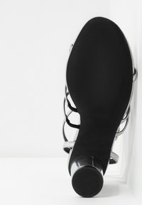 Sandale argentée à talons hauts avec une surface lisse et brillante. Présente des bandes entrecroisées et une semelle noire texturée. Design à bout rond.