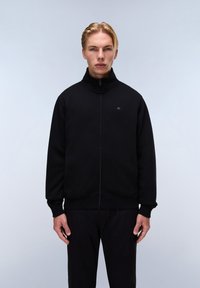 Černá mikina na zip s vysokým límcem, obsahující malé logo na hrudi a žebrované manžety a spodní lem. Materiál vypadá měkce a má texturovaný povrch.