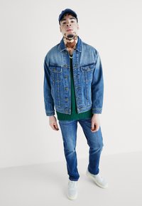 Jeans jakna svijetlog pranja, zelena rebrasta trenerka ispod, plave traperice i svijetle tenisice. Kapa u tamnoplavoj boji; ležerna odjeća.