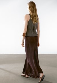 Grijze mouwloze top met een cinched taille, gecombineerd met een lange, bruine, vloeiende rok; draagt zwarte flip-flops en bruine armbanden.