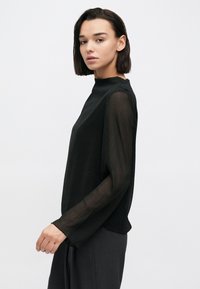 Μαύρο τοπ με μακριά, διαφανή μανίκια και ψηλό λαιμό, κατασκευασμένο από ελαφρύ ύφασμα. Απλός σχεδιασμός με άνετη εφαρμογή, συνδυασμένο με γκρι παντελόνι.