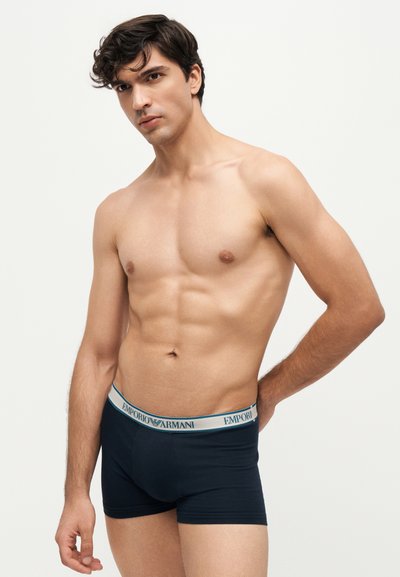 Γυμνόστηθος νεαρός άνδρας με σκούρα boxers Emporio Armani που στέκεται μπροστά σε απλό ανοιχτόχρωμο φόντο.