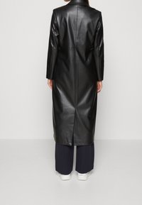 Vrouw in een lange zwarte leren jas, marineblauwe gestreepte broek en witte sneakers, staand met haar gezicht naar een effen grijze achtergrond.