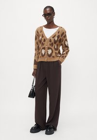 Brun leopardprikket cardigan med V-hals og knaplukning, parret med løse brune bukser og kraftige sorte sko. Sort håndtaske bemærket.