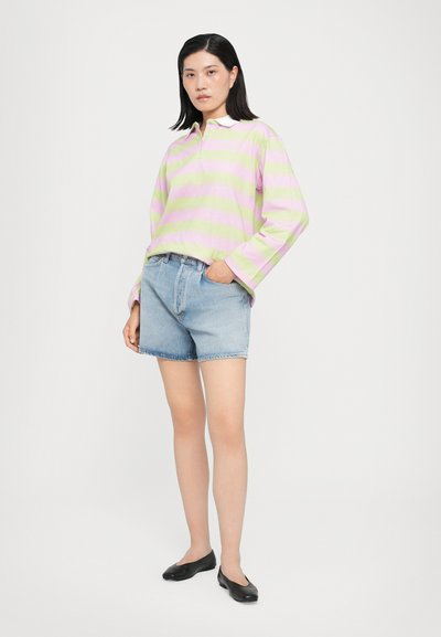 Polo s dolgimi rokavi v svetlo zeleni in roza črtah, v kombinaciji z denim kratkimi hlačami. Model nosi črne balerinke, kar prikazuje sproščen in neformalen videz.
