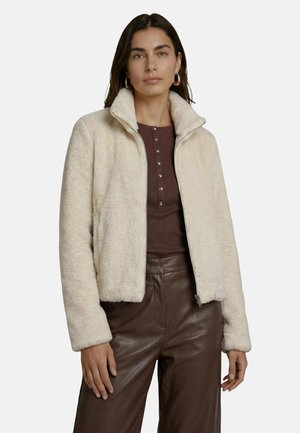 Femme portant des boucles d’oreilles créoles en or, une veste polaire crème sur un haut marron boutonné, et un pantalon en cuir marron foncé sur un fond uni.