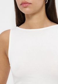 Femme portant un haut blanc sans manches avec des boucles d'oreilles créoles argentées, montrant le cou, l'épaule et les lèvres sur un fond neutre.
