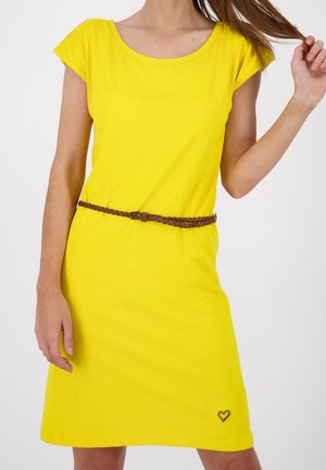 Frau trägt ein leuchtend gelbes Kurzarmkleid mit einem dünnen braunen geflochtenen Gürtel und einem kleinen Herz-Detail nahe dem Saum.