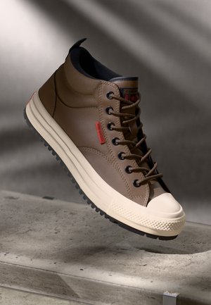 Chaussure mi-haute en cuir marron avec des lacets noirs, semelle en caoutchouc légère et capuchon de orteil texturé. Étiquette de marque rouge sur le côté.