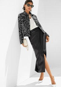 Frau trägt eine schwarz-weiß paisley-geblümte Jacke, eine weiße Bluse, einen schwarzen Midirock mit Schlitz, weiße High Heels, Sonnenbrille und hält eine kleine beigefarbene Handtasche.