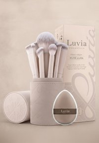 Sept pinceaux de maquillage vegan aux manches clairs dans un porte-pinceaux cylindrique texturé, une éponge en forme de goutte d'eau, et des emballages de la marque Luvia Cosmetics.