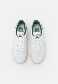 Beli športni copati z zelenimi detajli, iz platna in usnja, ravne vezalke, okrogla konica, podložen ovratnik in Lacoste logo na ježku.