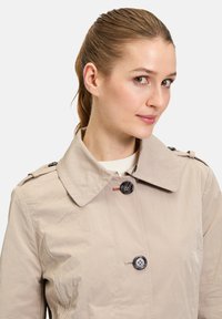 Hellbeige Trenchcoat mit weitem Kragen, schwarzen Knöpfen und Schulterklappen. Hergestellt aus einem glatten, leicht knitterigen Stoff.