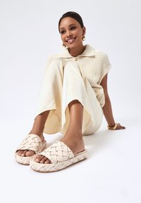 Geweven slider-sandalen in lichtbeige, met een dikke zool en open teendesign. Soepele textuur met een horizontaal gevlochten patroon.