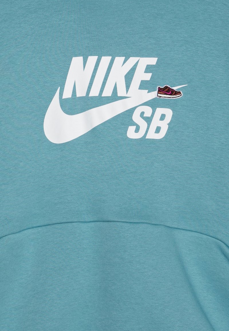 Türkisfarbener Sweatshirt mit einem großen weißen "NIKE SB"-Logo und einem gestickten Sneaker-Detail in Braun und Lila. Weiches, strukturiertes Material.