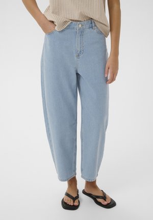 Model kannab helesiniseid, kõrge vöökohaga lahtisi teksapükse, beežikat triibulist toppi, mis on pükstesse sätitud, ja musti plätusid, seistes käed lõdvalt.