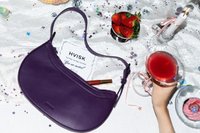 Fialová kožená taška cez rameno s oblým dizajnom a zipsom. Vedľa sú jahody, koktail, pozvánka na večierok a rozhádzané dekorácie.