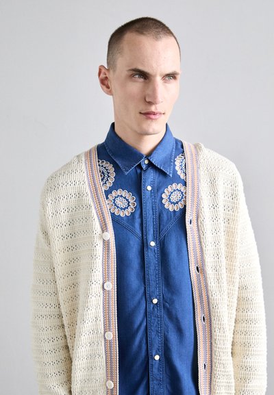 Gemütliche, cremefarbene Strickjacke mit offenem Muster und pastellfarbigen Akzenten, kombiniert mit einem blauen Denimhemd, das mit floralen Stickereien verziert ist.