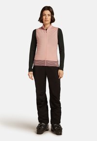 Donna che indossa un gilet rosa senza maniche con zip sopra una maglietta nera a maniche lunghe, pantaloni neri e stivali da sci neri, in piedi su uno sfondo bianco.