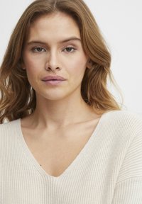 Pull en maille beige clair avec un col en V, présentant un motif texturé et une coupe ample. Le tissu semble doux et légèrement extensible.