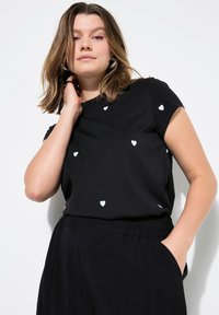 Černé tričko s krátkým rukávem, které má malé bílé motivy srdcí rozptýlené po přední části; měkká tkanina a volný střih. V kombinaci s černými kalhotami.